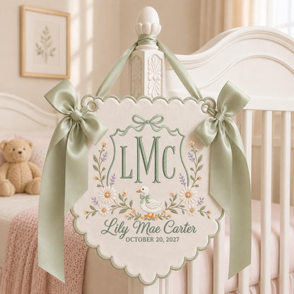 May include: A cream-colored fabric wall hanging with scalloped edges, featuring sage green embroidery of the initials LMC, a duck, and floral accents. It includes the name Lily Mae Carter and the date OCTOBER 20, 2027, with sage green ribbon bows.