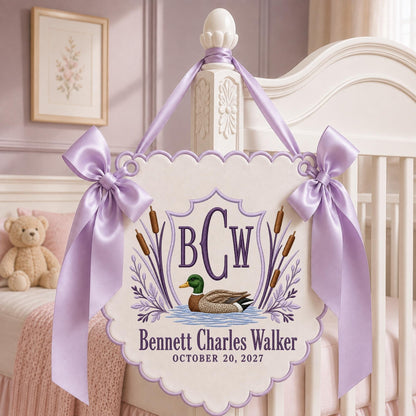 May include: A cream-colored scalloped wall hanging featuring an embroidered mallard duck, cattails, and the monogram BCW. The text reads Bennett Charles Walker and October 20, 2027. It is decorated with two lavender satin bows and matching ribbon.