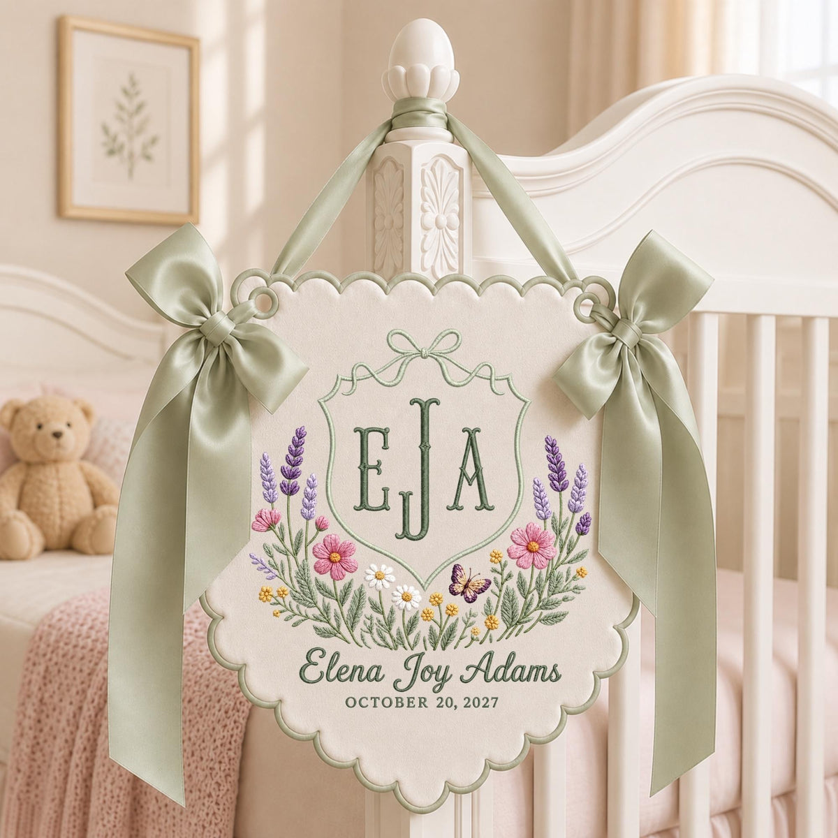 May include: A cream-colored fabric wall hanging with scalloped edges, featuring embroidered lavender, pink, and yellow flowers, a butterfly, and the monogram EJA. Below, the text reads Elena Joy Adams and OCTOBER 20, 2027, with sage green ribbons.