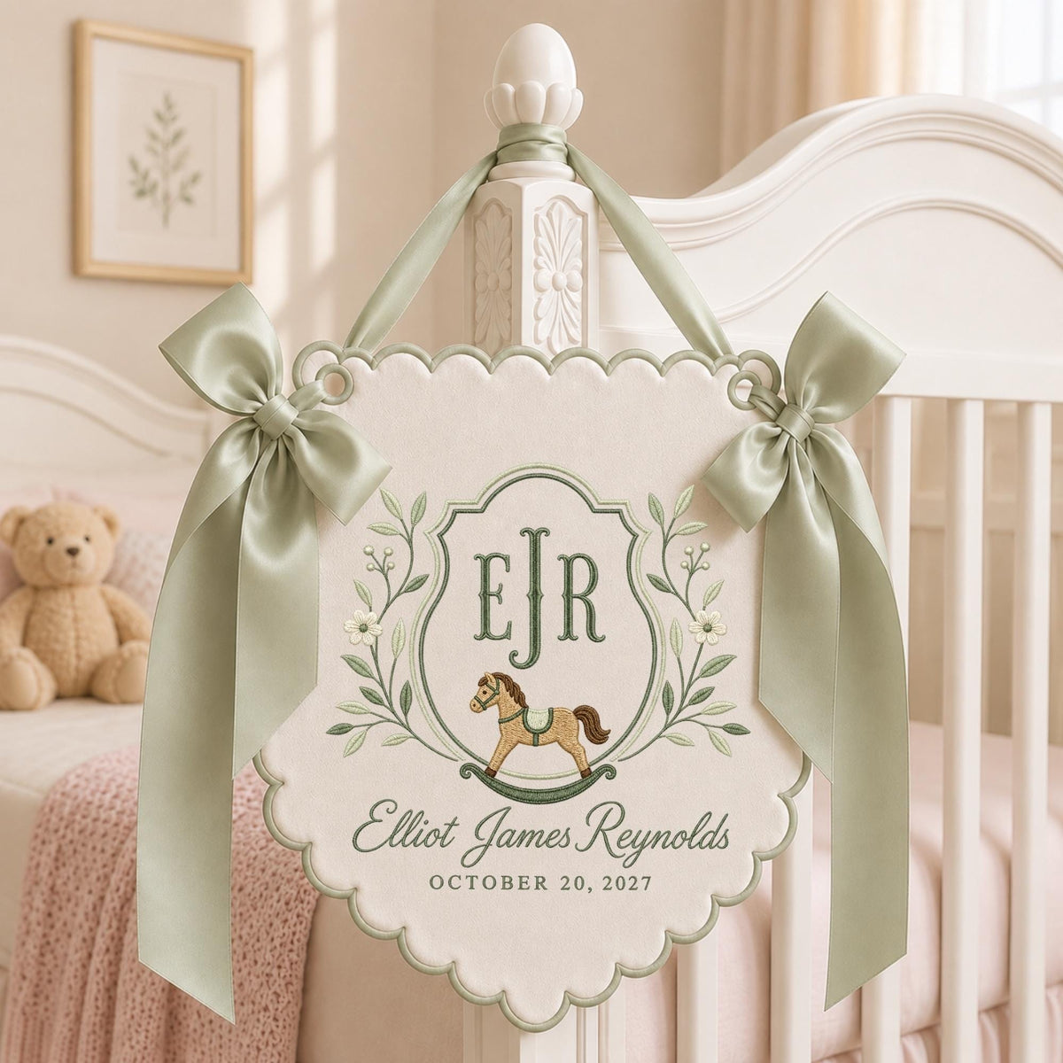 May include: A cream-colored fabric wall hanging with scalloped edges, featuring sage green satin bows on each side. The embroidery includes the monogram EJR, a rocking horse, the name Elliot James Reynolds, and the date OCTOBER 20, 2027.