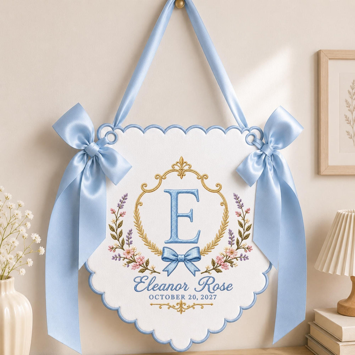 May include: A white scalloped wall hanging banner with light blue satin ribbon bows on each side and a matching hanging ribbon. The center features a gold embroidered frame, a large blue letter E, floral accents, and the text Eleanor Rose OCTOBER 20, 2027.