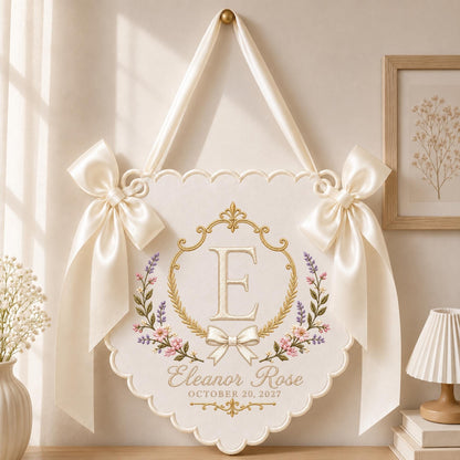May include: A cream-colored fabric wall hanging with a scalloped edge, featuring an embroidered gold frame, the letter E, and floral accents. It includes the text Eleanor Rose and OCTOBER 20, 2027, finished with two large satin ribbon bows.
