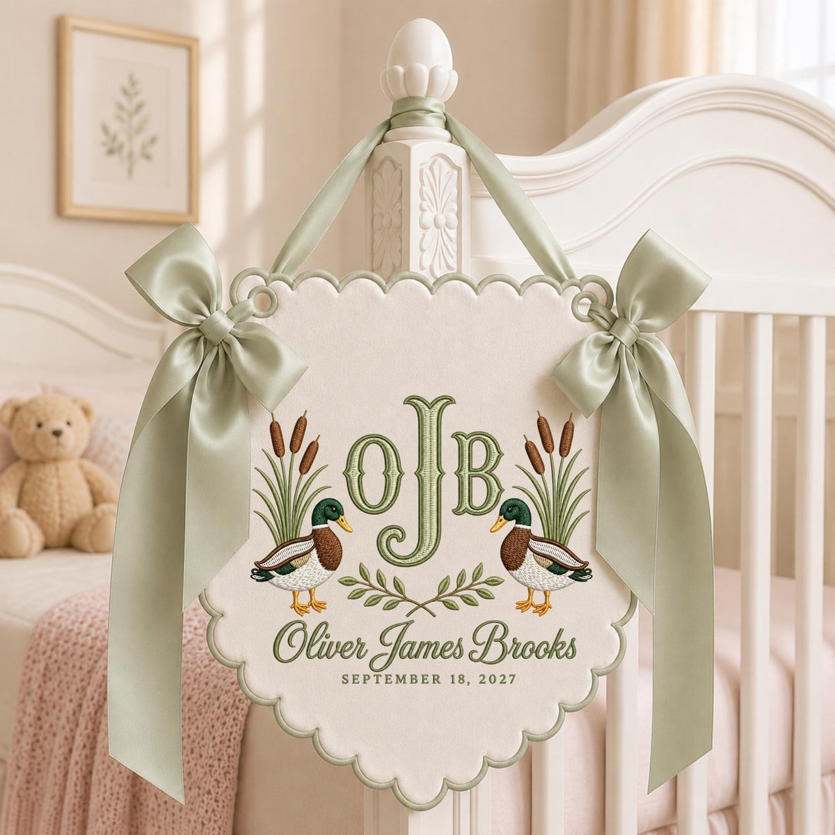 May include: A cream-colored scalloped wall hanging featuring embroidered mallard ducks, cattails, and the monogram OJB. The text reads Oliver James Brooks and September 18, 2027. It is decorated with two sage green satin ribbons tied in bows.
