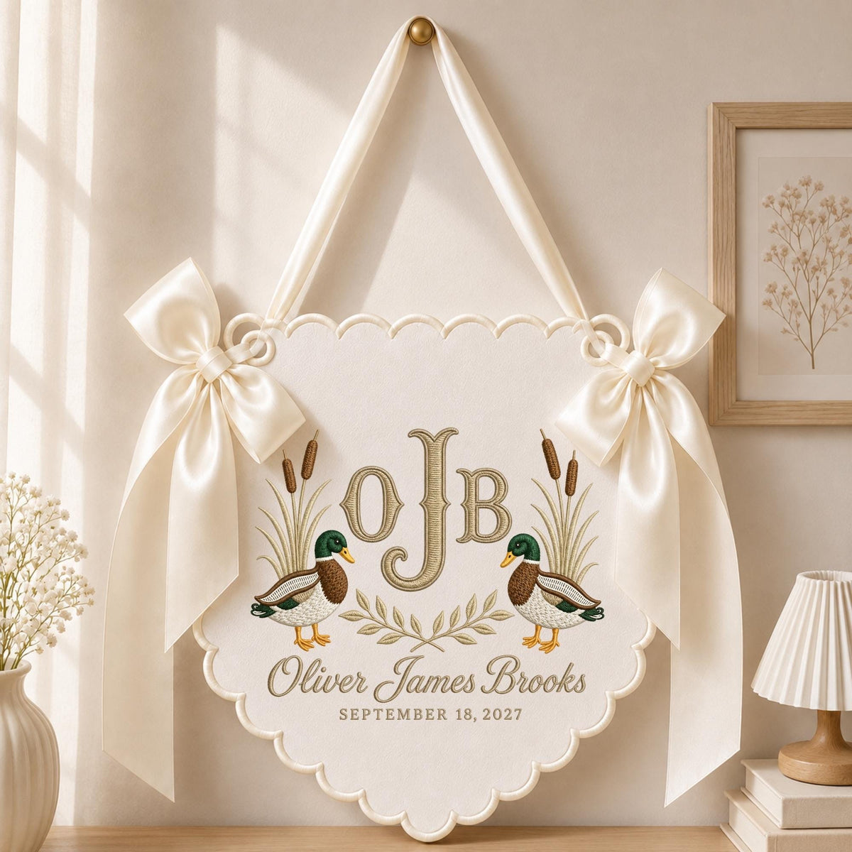 May include: A cream-colored scalloped wall hanging featuring two embroidered mallard ducks, cattails, and a laurel branch. The center displays the monogram OJB, the name Oliver James Brooks, and the date SEPTEMBER 18, 2027. Two satin bows hang from the top.