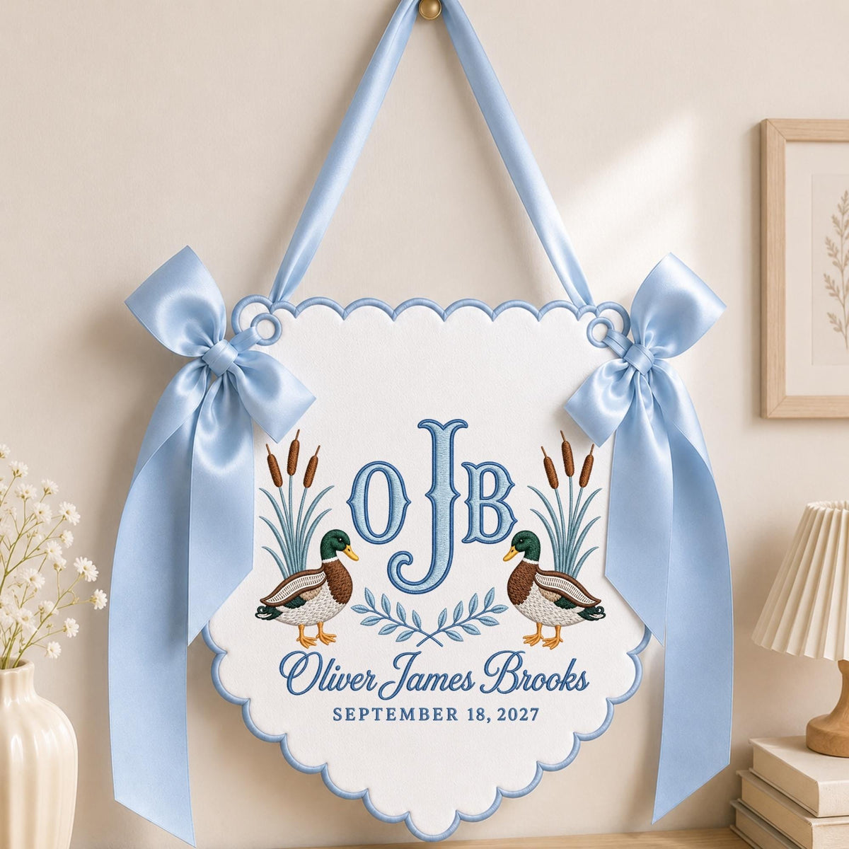 May include: A white scalloped wall hanging featuring two mallard ducks, cattails, and the embroidered monogram OJB. Below the monogram, the text reads Oliver James Brooks and SEPTEMBER 18, 2027. The sign is accented with light blue satin ribbons.