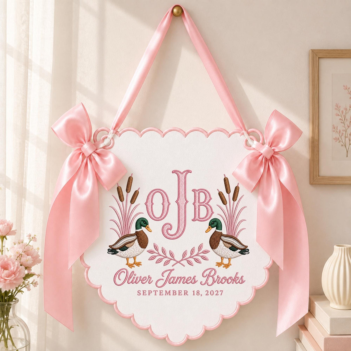 May include: A white scalloped wall hanging featuring two mallard ducks, pink embroidered cattails, and the monogram OJB. Below the ducks, the text reads Oliver James Brooks and September 18, 2027. The sign is accented with two large pink satin bows.