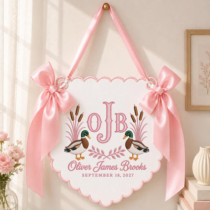May include: A white scalloped wall hanging featuring two mallard ducks, pink embroidered cattails, and the monogram OJB. Below the ducks, the text reads Oliver James Brooks and September 18, 2027. The sign is accented with two large pink satin bows.