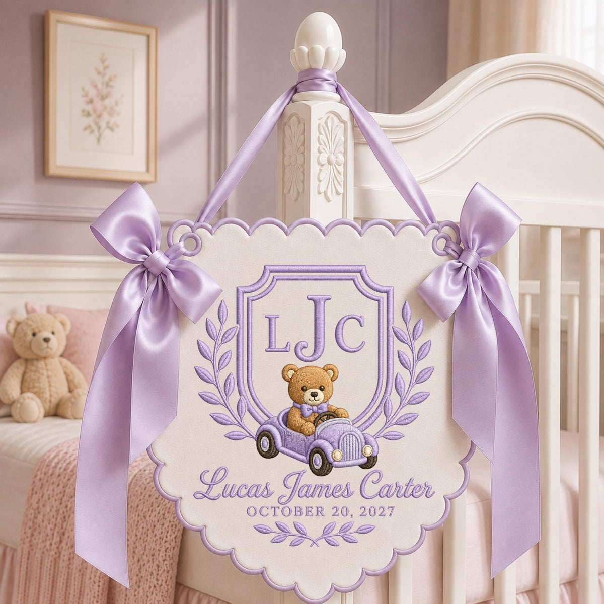 May include: A cream-colored fabric wall hanging with scalloped edges, featuring lavender embroidery of a teddy bear in a car, the initials LJC, the name Lucas James Carter, and the date OCTOBER 20, 2027, accented by two large lavender satin bows.