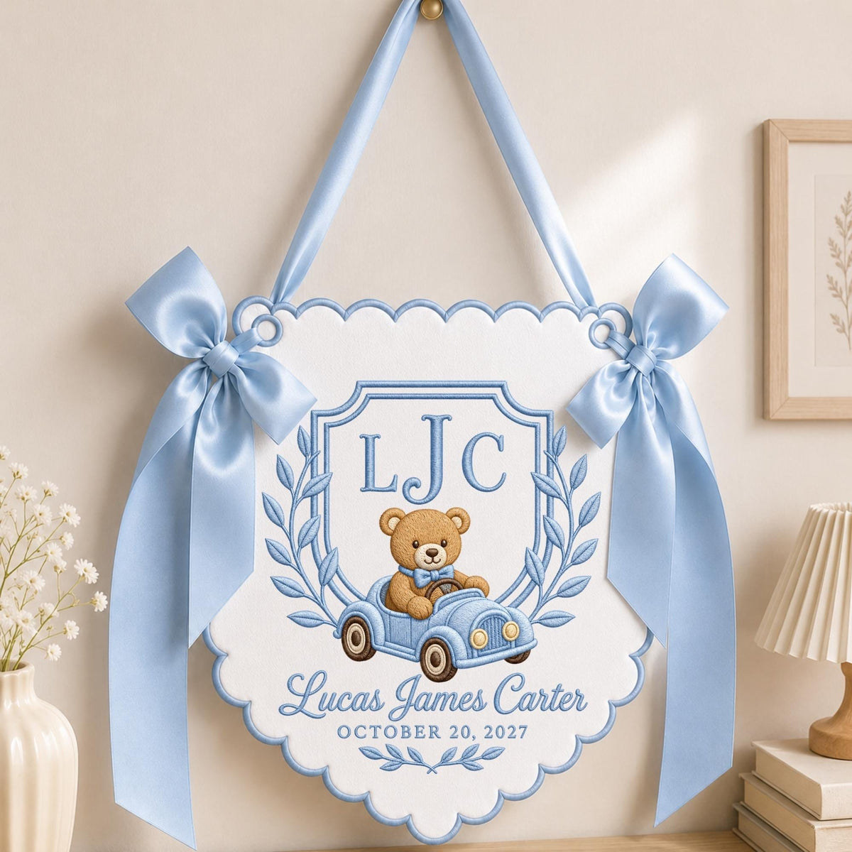May include: A white scalloped wall hanging with light blue satin ribbon bows and trim. It features an embroidered crest with the initials LJC, a brown teddy bear in a light blue toy car, and the text Lucas James Carter OCTOBER 20, 2027.
