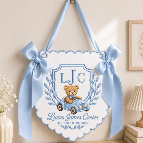 May include: A white scalloped wall hanging with light blue satin ribbon bows and trim. It features an embroidered crest with the initials LJC, a brown teddy bear in a light blue toy car, and the text Lucas James Carter OCTOBER 20, 2027.