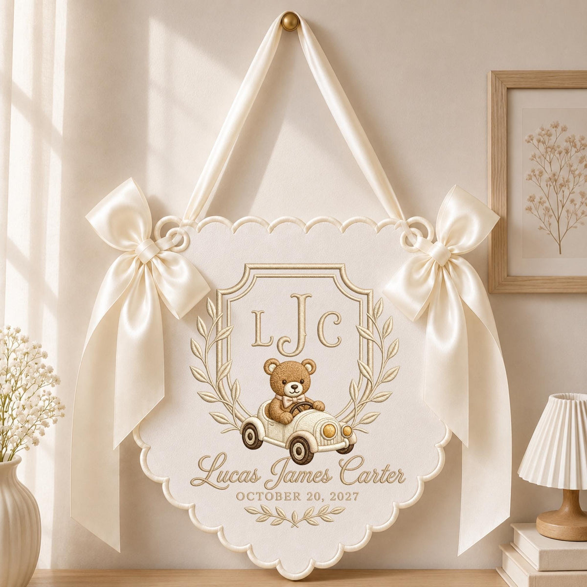 May include: A cream-colored fabric wall hanging with scalloped edges, featuring two large satin bows on the sides and a ribbon for hanging. The design includes a teddy bear in a vintage car, the initials LJC, and the text Lucas James Carter October 20, 2027.