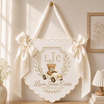 May include: A cream-colored fabric wall hanging with scalloped edges, featuring two large satin bows on the sides and a ribbon for hanging. The design includes a teddy bear in a vintage car, the initials LJC, and the text Lucas James Carter October 20, 2027.