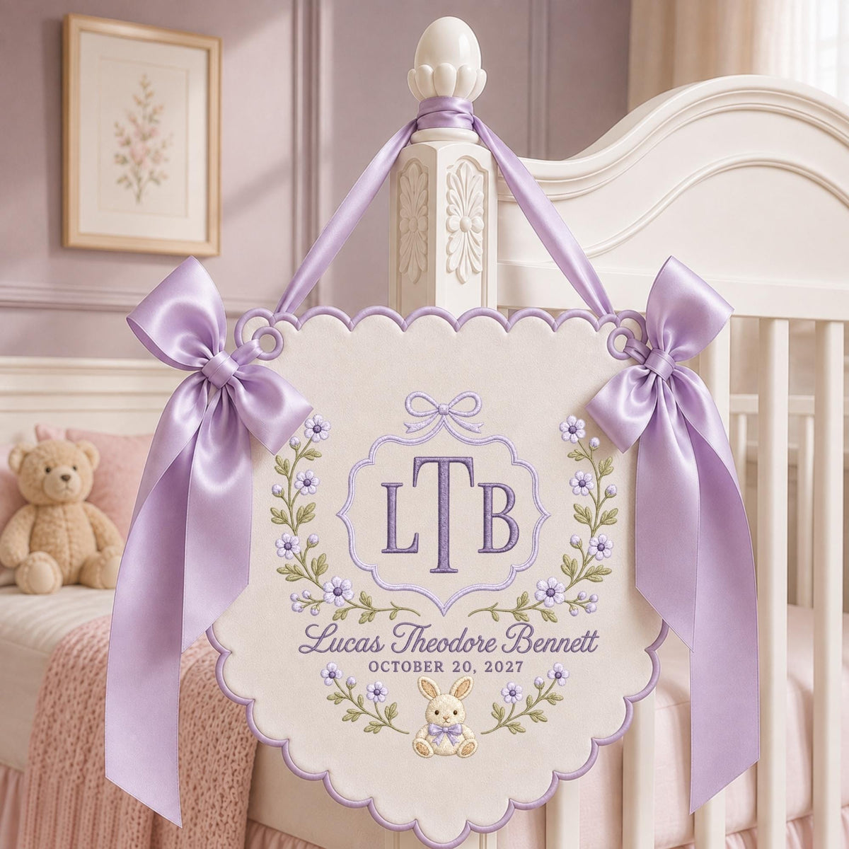May include: A cream-colored fabric wall hanging with scalloped edges, featuring purple embroidery of the monogram LTB, the name Lucas Theodore Bennett, the date October 20, 2027, and a small bunny. It is adorned with two large lavender satin bows.