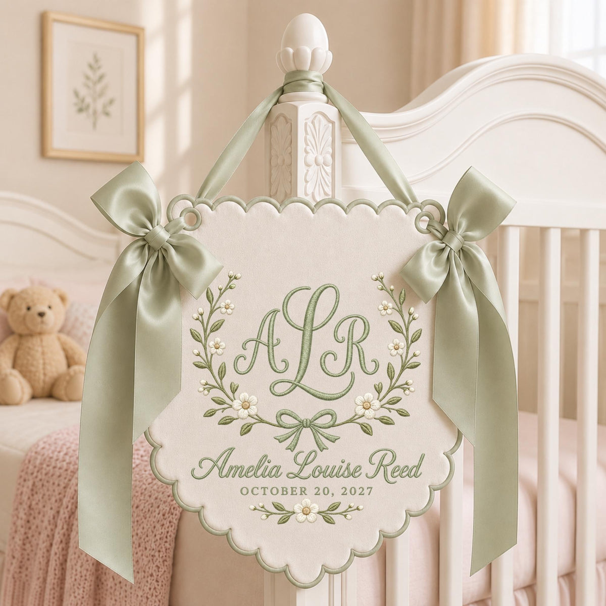 May include: A cream-colored scalloped wall hanging featuring sage green embroidery of the initials ALR, a floral wreath, and the text Amelia Louise Reed October 20, 2027. The banner is accented with two sage green satin ribbons tied at the top.