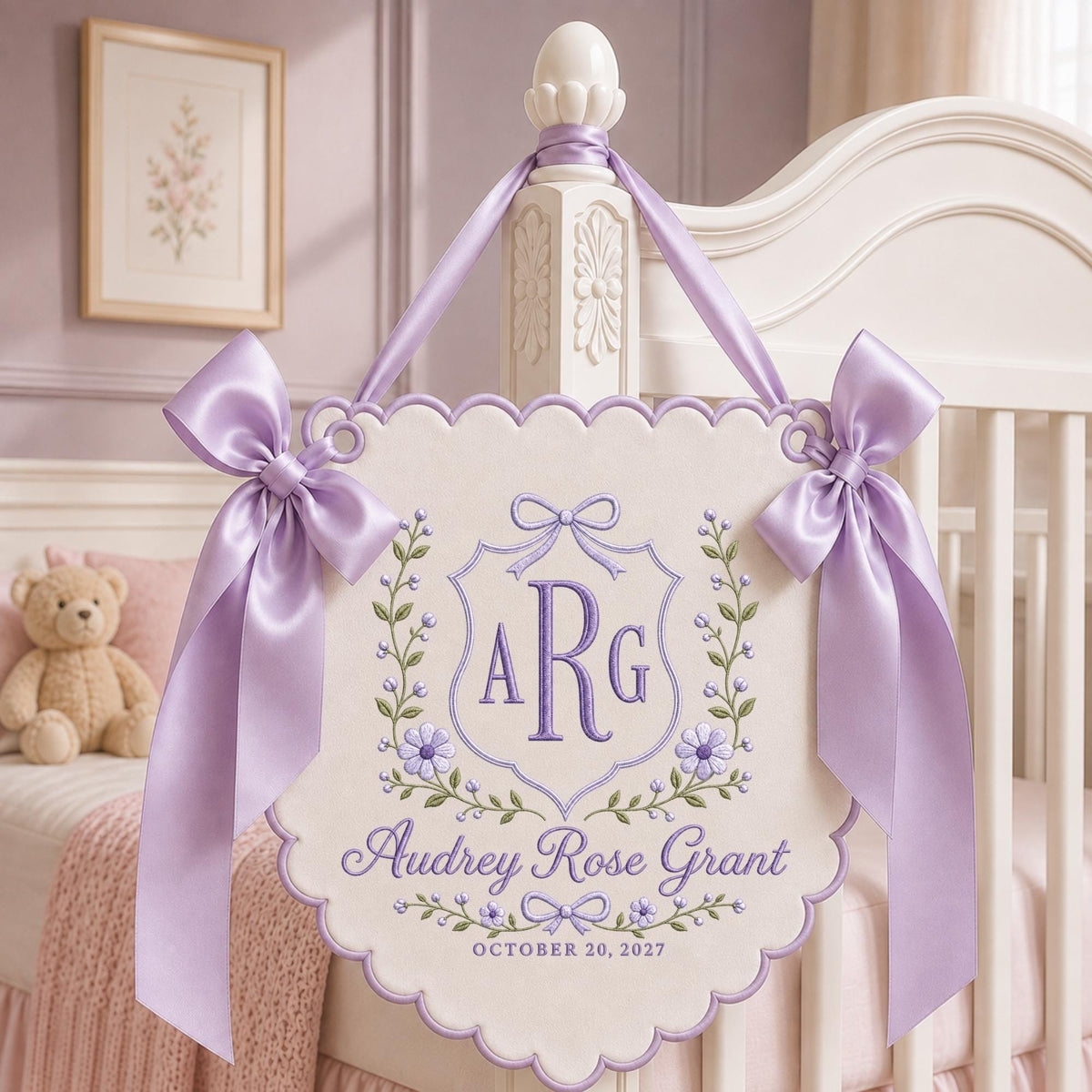 May include: A cream-colored scalloped wall hanging featuring purple embroidery of the monogram ARG, the name Audrey Rose Grant, and the date October 20, 2027. The piece is adorned with floral vines and two large lavender satin ribbon bows.