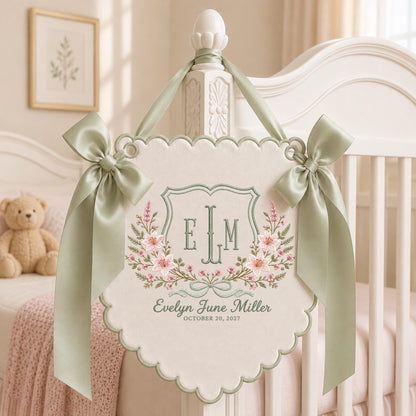 May include: A cream-colored scalloped wall hanging featuring sage green satin bows on each side. The embroidery includes a central shield with the monogram E L M, floral accents in pink and green, and the text Evelyn June Miller October 20, 2027.