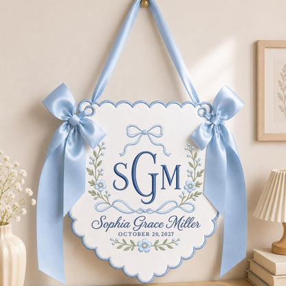 May include: A white scalloped wall hanging featuring blue embroidered floral vines, a central monogram SGM, and the text Sophia Grace Miller OCTOBER 20, 2027. The piece is suspended by a light blue satin ribbon and adorned with two matching side bows.