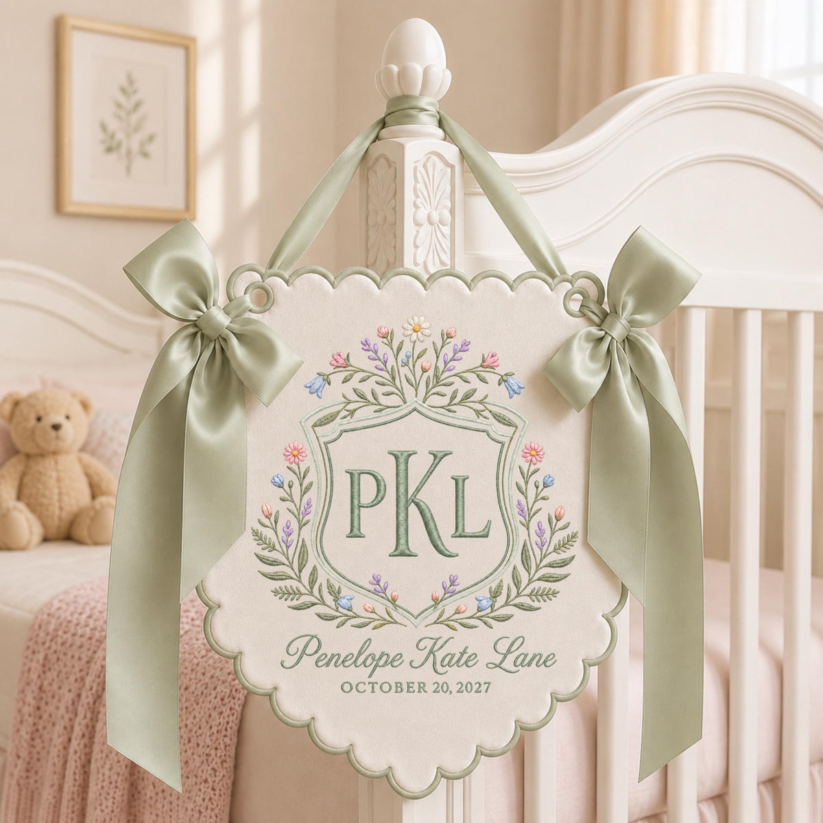 May include: A cream-colored scalloped wall hanging featuring an embroidered crest with the initials PKL, surrounded by floral motifs in pink, blue, and green. The text reads Penelope Kate Lane and OCTOBER 20, 2027, accented by sage green satin bows.