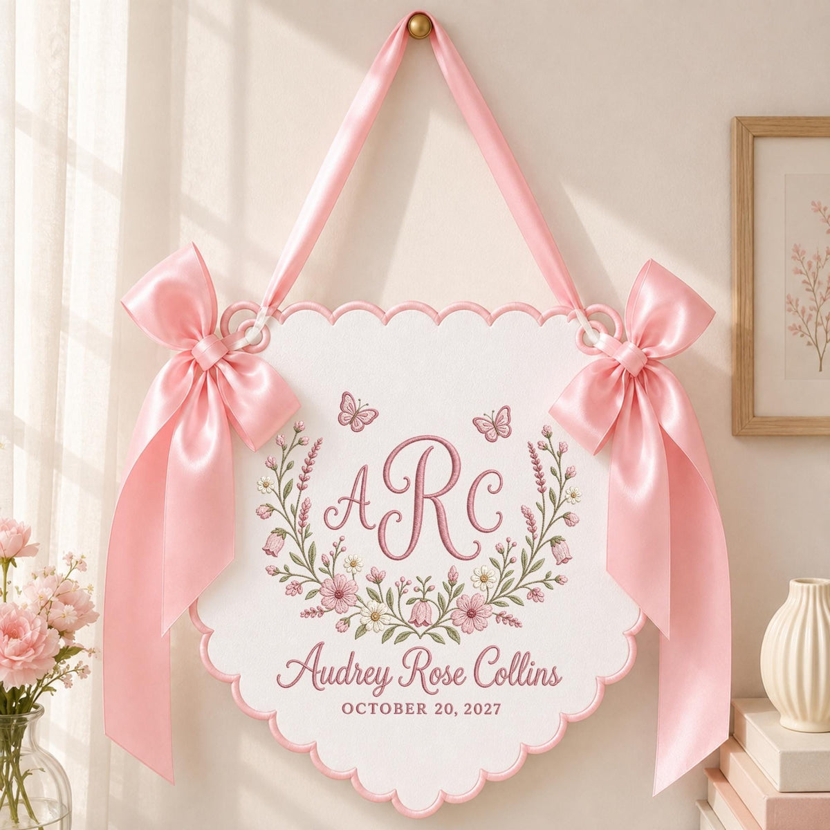 May include: A white scalloped wall hanging featuring a pink embroidered monogram ARC, a floral wreath, two butterflies, and the text Audrey Rose Collins OCTOBER 20, 2027. The sign is suspended by a pink satin ribbon and adorned with two large pink bows.