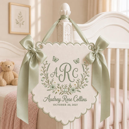May include: A cream-colored fabric wall hanging with scalloped edges, featuring sage green embroidery of the initials ARC, a floral wreath, two butterflies, and the text Audrey Rose Collins and OCTOBER 20, 2027, accented by two sage green satin bows.