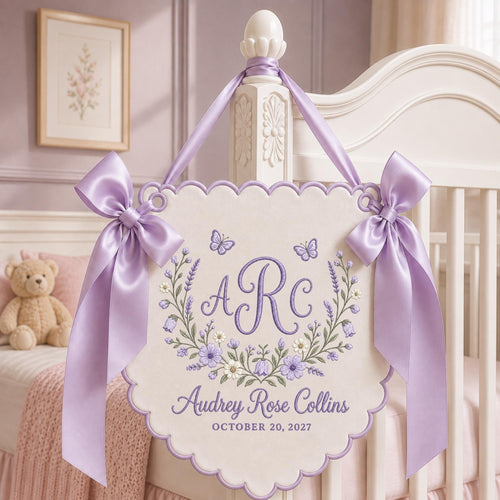 May include: A cream-colored scalloped wall hanging featuring purple embroidery of the monogram ARC, a floral wreath, two butterflies, and the text Audrey Rose Collins and OCTOBER 20, 2027. It is adorned with two large lavender satin bows.