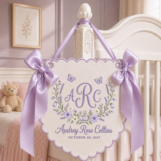 May include: A cream-colored scalloped wall hanging featuring purple embroidery of the monogram ARC, a floral wreath, two butterflies, and the text Audrey Rose Collins and OCTOBER 20, 2027. It is adorned with two large lavender satin bows.