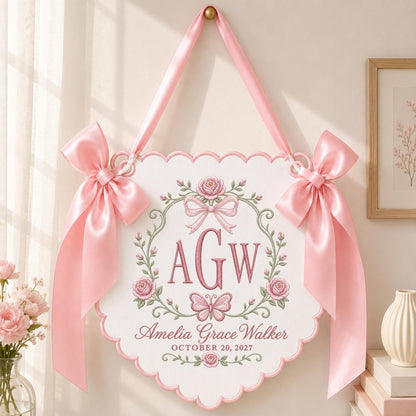 May include: A white scalloped wall hanging featuring the pink embroidered monogram AGW, a butterfly, and rose floral motifs. The text reads Amelia Grace Walker and OCTOBER 20, 2027. It is suspended by a pink satin ribbon with large pink bows on each side.
