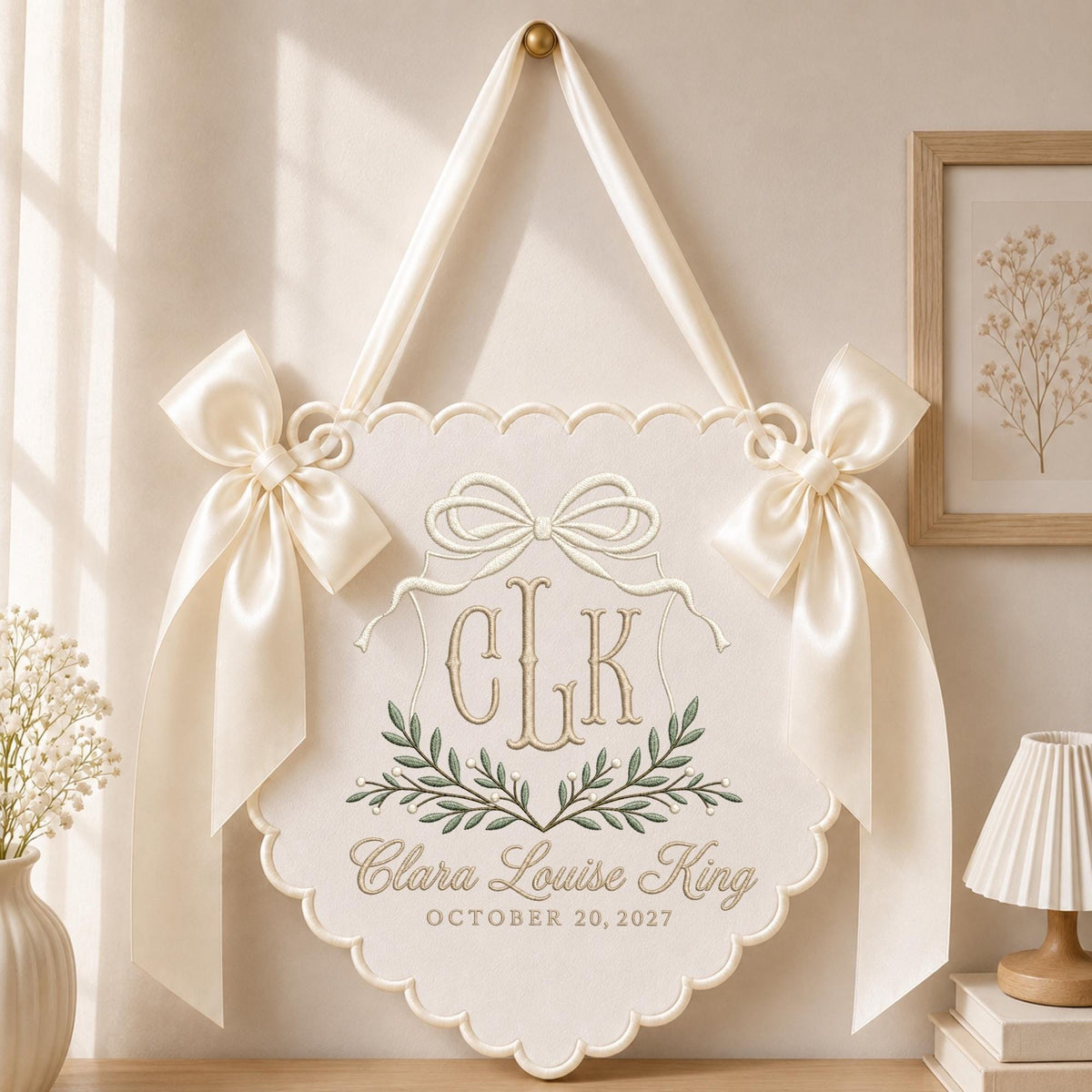 May include: A cream-colored fabric wall hanging with a scalloped edge, featuring an embroidered monogram CLK, a bow, and green leaf motifs. The text Clara Louise King and OCTOBER 20, 2027 is embroidered below. Two satin ribbons are tied at the top sides.