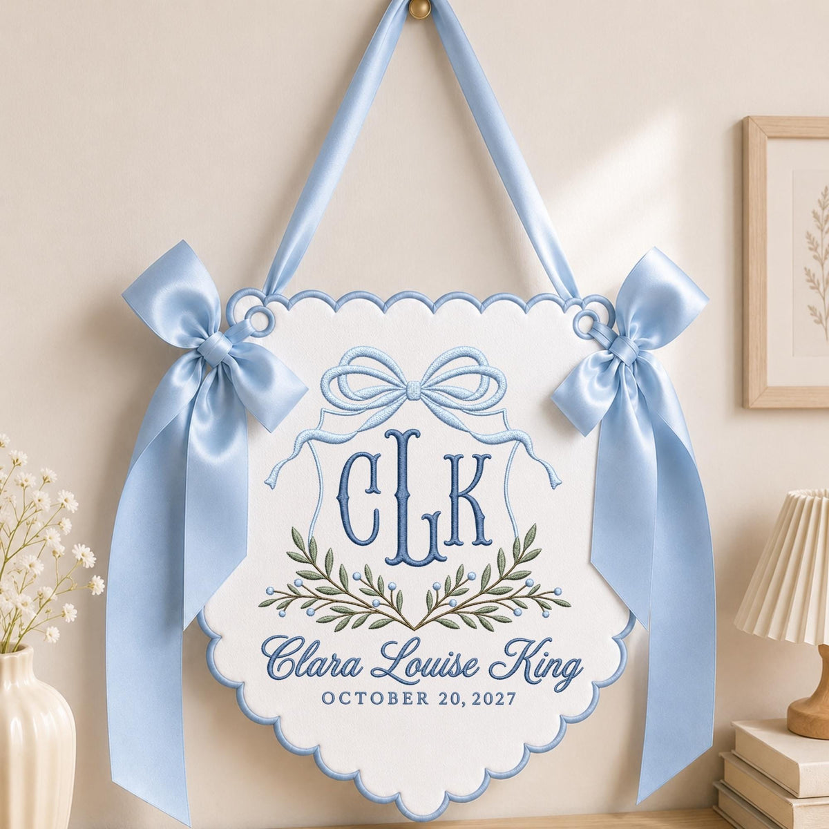 May include: A white scalloped wall hanging featuring blue embroidered details, including a central bow, the monogram CLK, and the text Clara Louise King October 20, 2027. The piece is accented with green leaf embroidery and light blue satin ribbon bows.