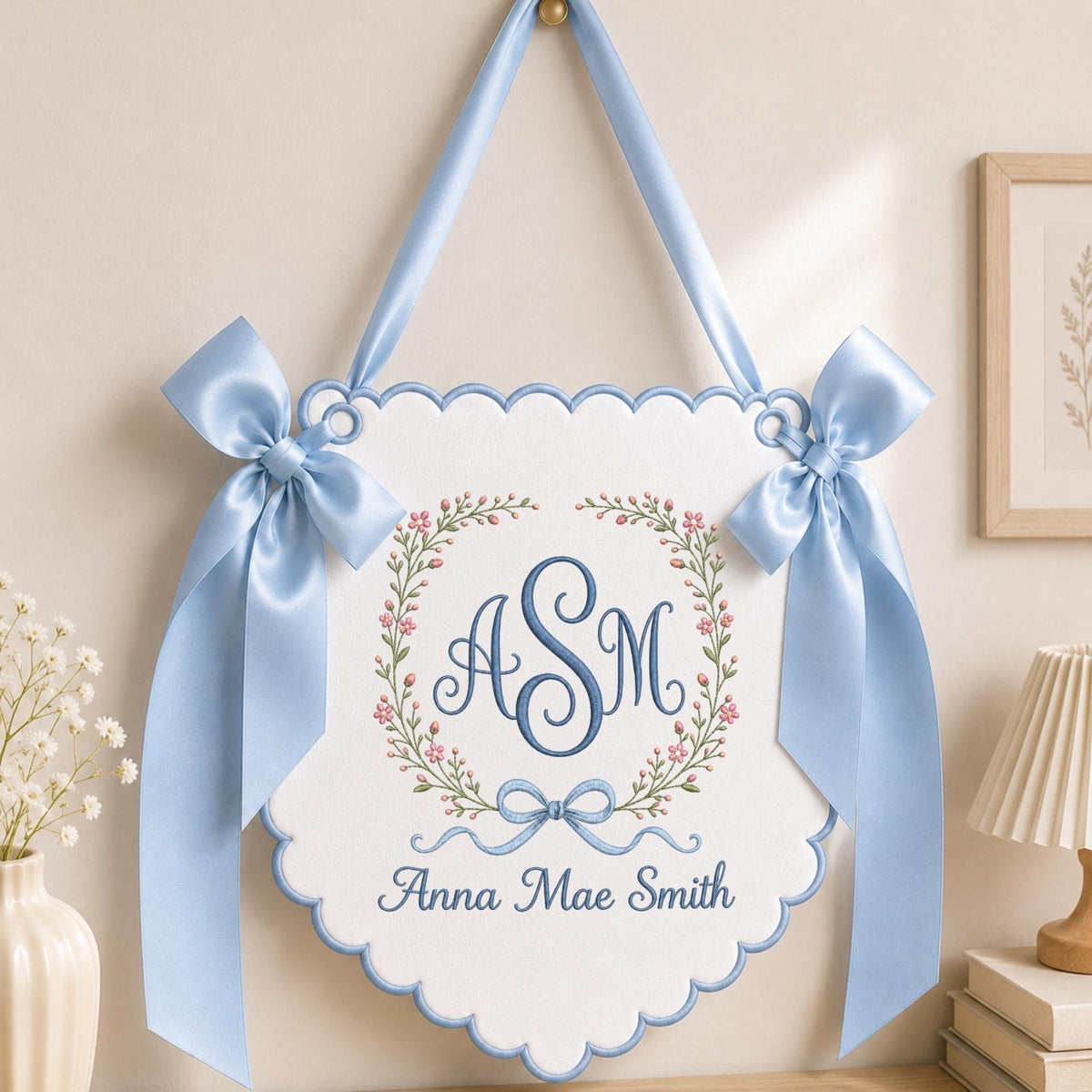 May include: A white scalloped wall hanging featuring an embroidered blue monogram with the letters ASM, surrounded by a floral wreath of green leaves and pink flowers. The piece includes the name Anna Mae Smith and two light blue satin bows.