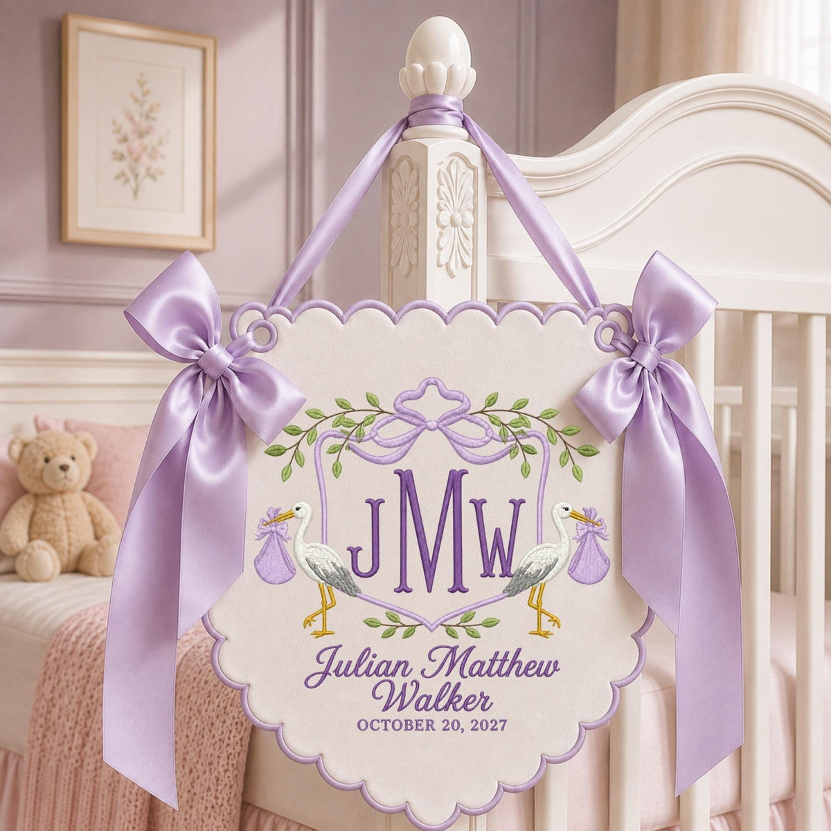 May include: A scalloped, off-white fabric wall hanging featuring purple embroidery of the initials JMW, two storks carrying purple bundles, and the text Julian Matthew Walker OCTOBER 20, 2027. It is adorned with two large lavender satin bows.