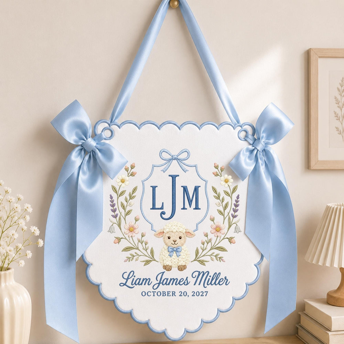 May include: A white scalloped wall hanging featuring a blue embroidered monogram LJM, a central illustration of a lamb with a blue bow, and the text Liam James Miller October 20, 2027. The piece is accented with two large light blue satin bows.