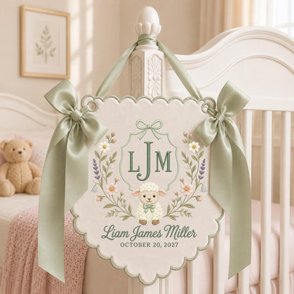 May include: A cream-colored scalloped wall hanging featuring an embroidered lamb, floral wreath, and the monogram LJM. The text reads Liam James Miller and October 20, 2027. It is accented with sage green satin ribbons tied at the top corners.
