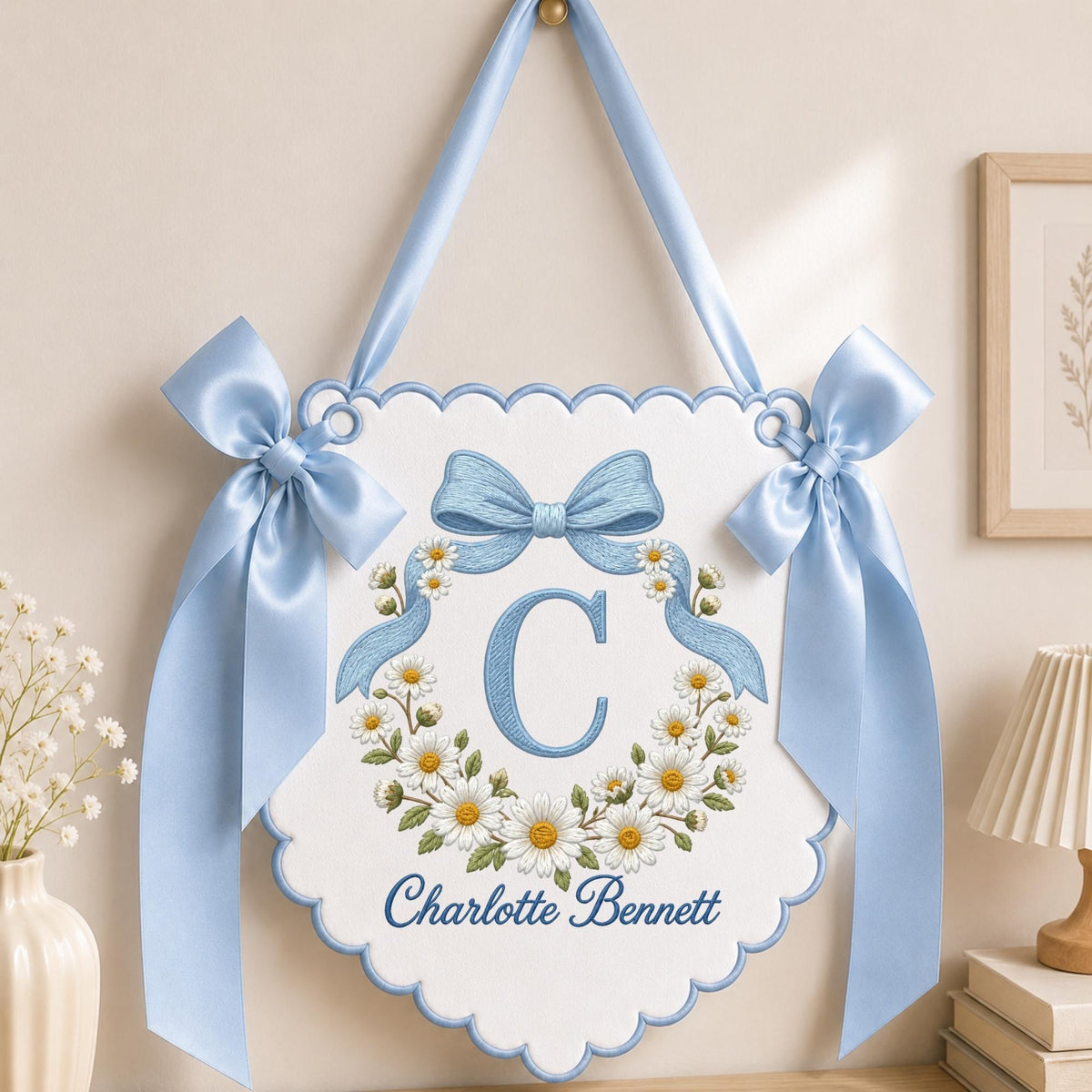 May include: A white scalloped wall hanging banner featuring a blue embroidered letter C surrounded by a daisy wreath. The banner includes the name Charlotte Bennett in blue script, accented by two large light blue satin bows and a hanging ribbon.