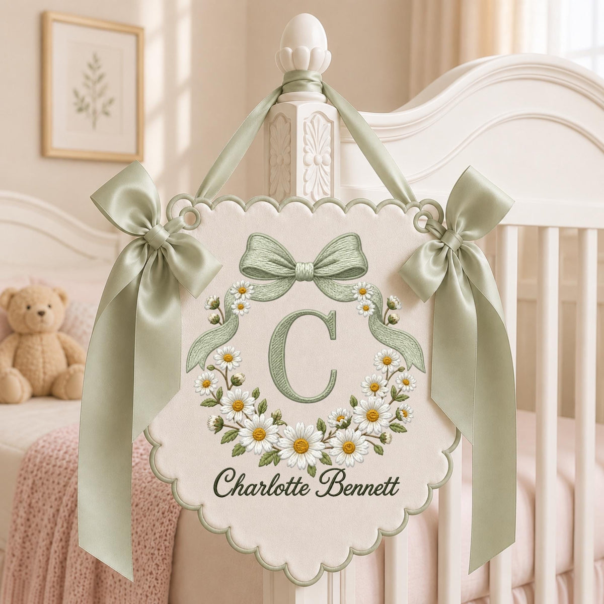May include: A cream-colored fabric wall hanging with a scalloped edge, featuring an embroidered sage green letter C surrounded by a wreath of white daisies. The banner is accented with two sage green satin bows and the name Charlotte Bennett.