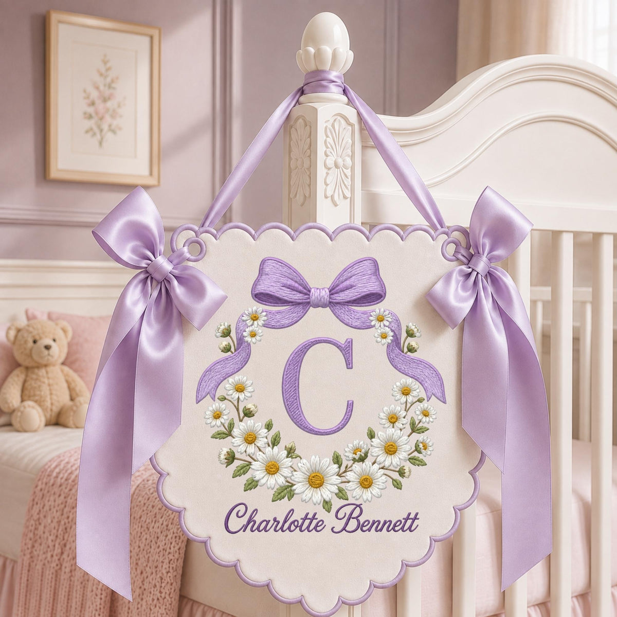 May include: A scalloped, cream-colored fabric wall hanging featuring a purple embroidered letter C surrounded by a daisy wreath and a purple bow. The name Charlotte Bennett is embroidered below in purple, with lavender satin ribbons on each side.