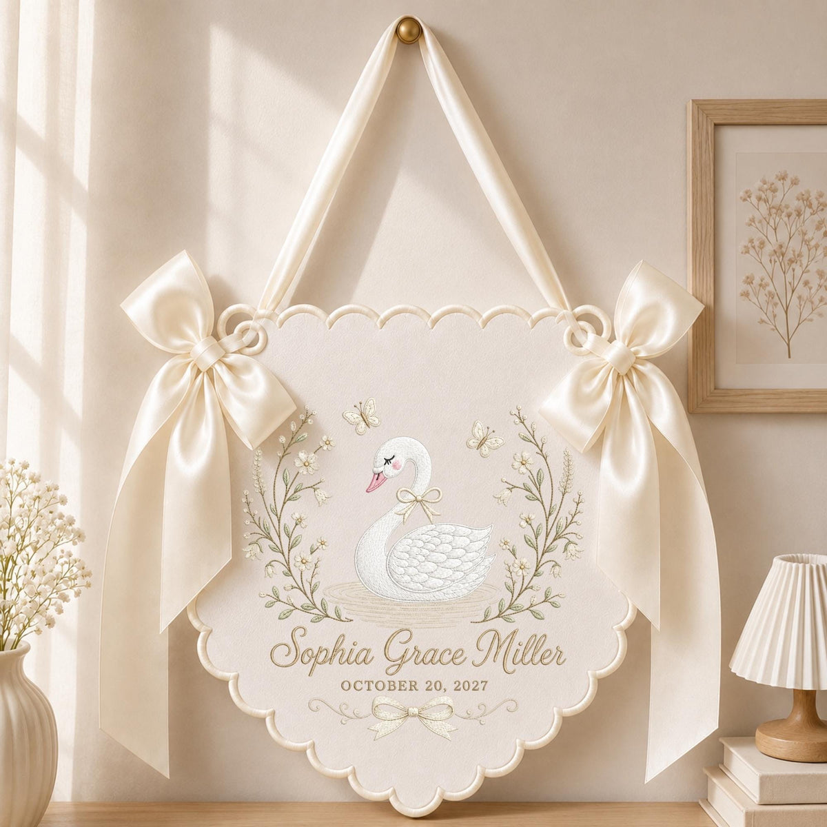 May include: A cream-colored scalloped wall hanging featuring a printed illustration of a white swan surrounded by floral vines and butterflies. The text reads Sophia Grace Miller and October 20, 2027. It is accented with two large satin bows.