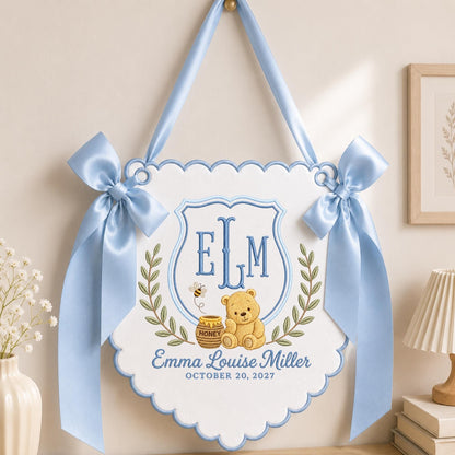 May include: A white scalloped wall hanging featuring light blue satin ribbon bows on each side. The center displays an embroidered blue shield with the letters ELM, a yellow bear, a honey pot, a bee, and the text Emma Louise Miller October 20, 2027.