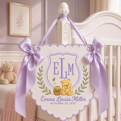 May include: A cream-colored scalloped wall hanging featuring purple embroidery of the monogram ELM, a teddy bear, a honey pot, and a bee. The text reads Emma Louise Miller and October 20, 2027. It is decorated with two large lavender satin bows.