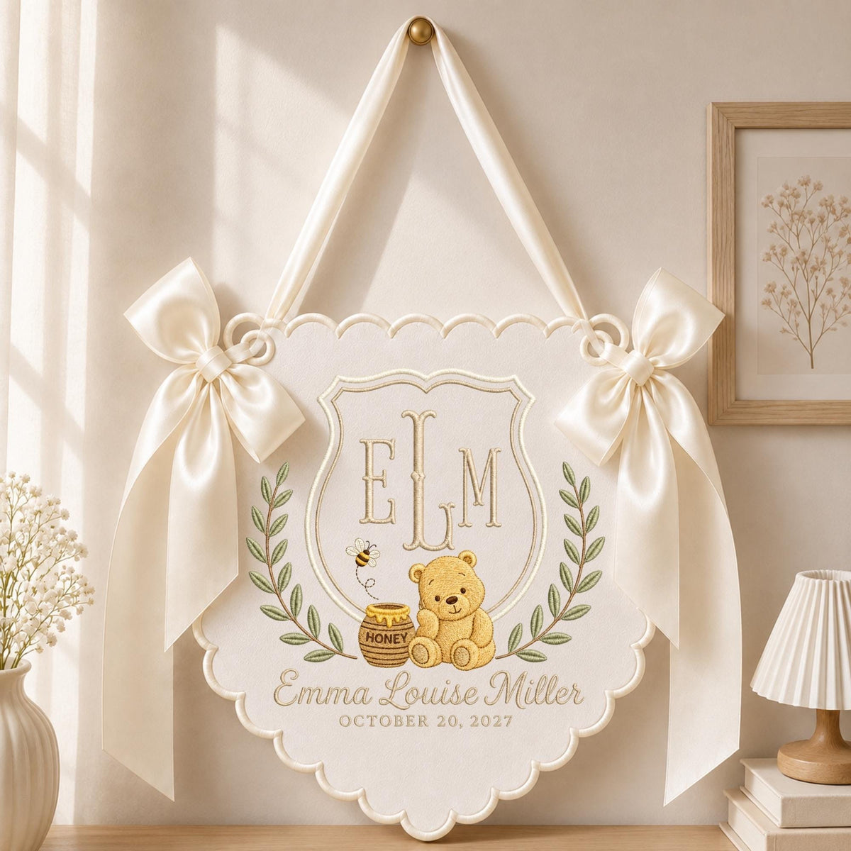 May include: A cream-colored scalloped wall hanging featuring an embroidered shield with the monogram ELM, a teddy bear, a honey pot, and a bee. The text reads Emma Louise Miller and October 20, 2027. It is accented with two large satin ribbons.