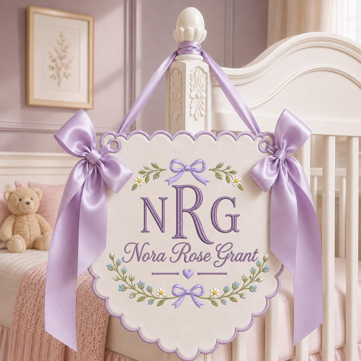 May include: A scalloped, off-white fabric wall hanging featuring purple embroidered text that reads NRG and Nora Rose Grant. The design includes floral motifs, small bows, and a heart. It is suspended by a lavender satin ribbon with large bows.