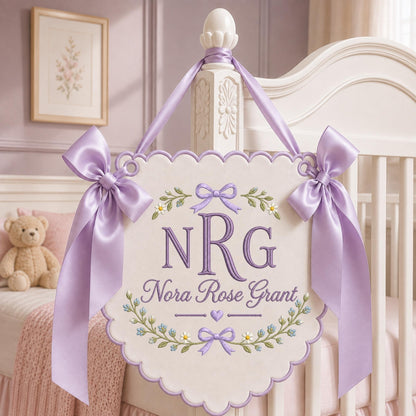 May include: A scalloped, off-white fabric wall hanging featuring purple embroidered text that reads NRG and Nora Rose Grant. The design includes floral motifs, small bows, and a heart. It is suspended by a lavender satin ribbon with large bows.