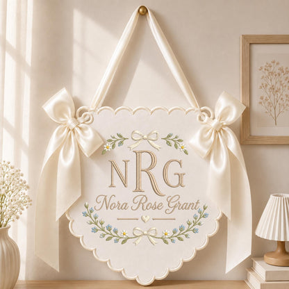 May include: A cream-colored scalloped wall hanging featuring the embroidered monogram NRG and the name Nora Rose Grant. The design includes floral vines, small blue flowers, and a central heart. Two large satin ribbons are tied at the top corners.
