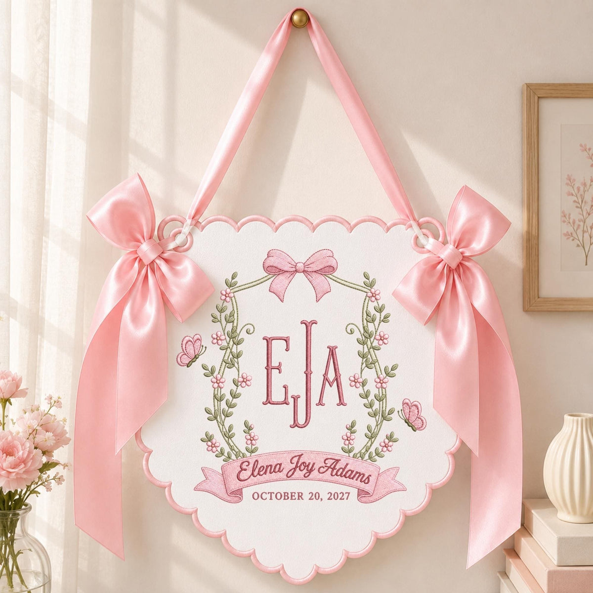 May include: A white scalloped wall hanging featuring pink satin bows on each side and a pink ribbon for hanging. The center displays the embroidered monogram EJA, a floral wreath, two butterflies, and the text Elena Joy Adams October 20, 2027.