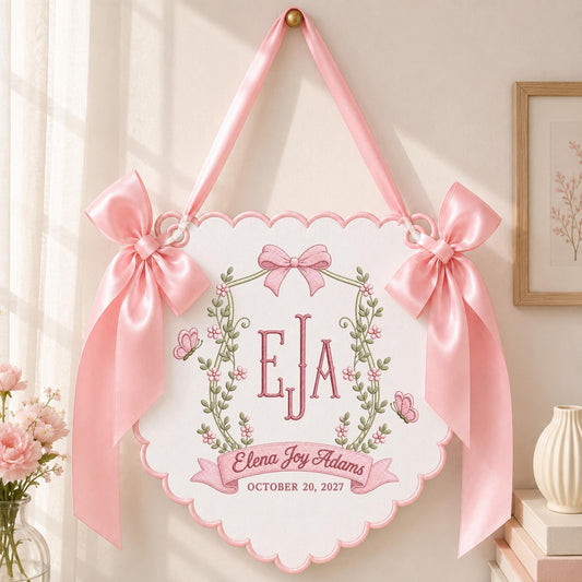 May include: A white scalloped wall hanging featuring pink satin bows on each side and a pink ribbon for hanging. The center displays the embroidered monogram EJA, a floral wreath, two butterflies, and the text Elena Joy Adams October 20, 2027.