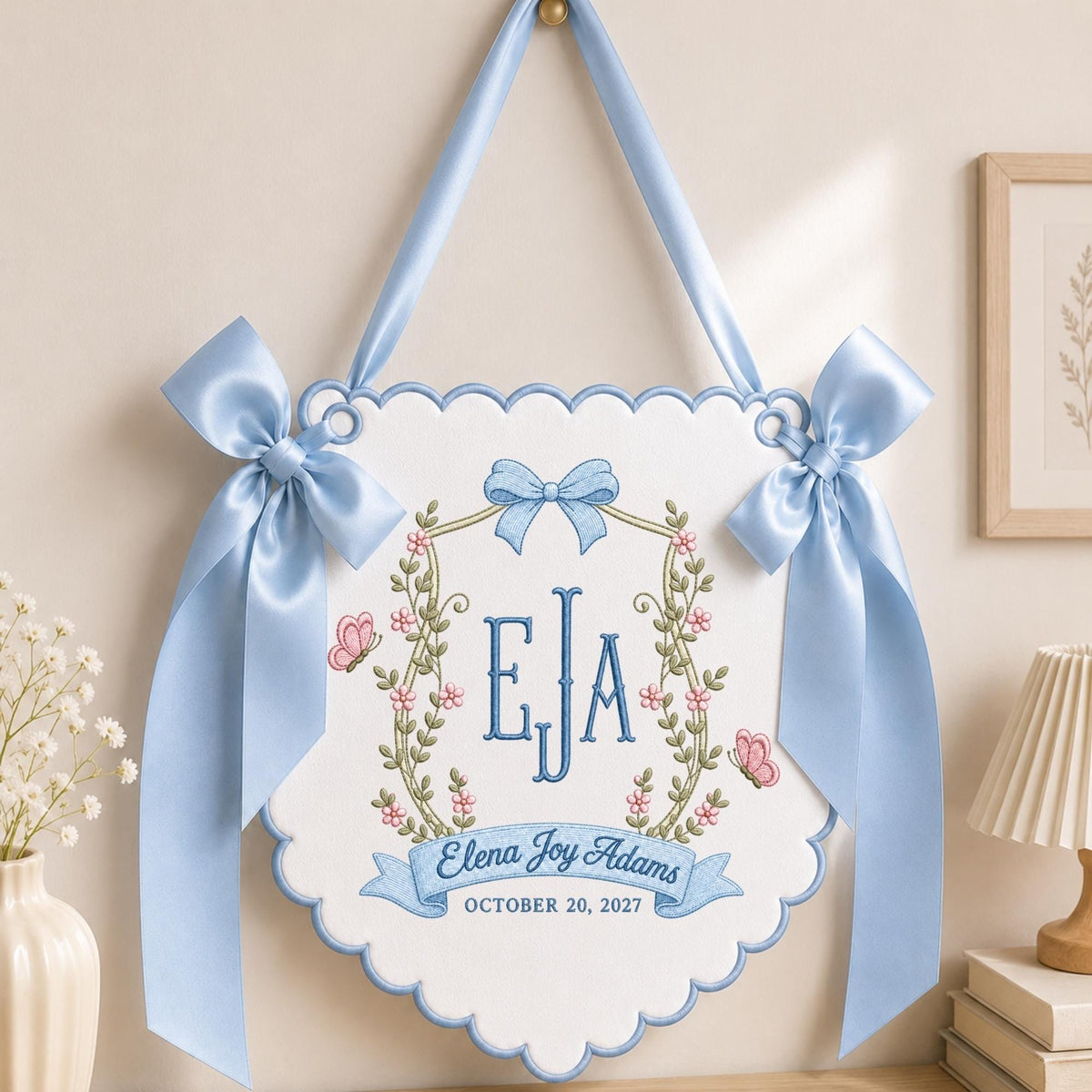 May include: A white scalloped fabric wall hanging featuring blue embroidered monogram letters EJA, a floral wreath with pink flowers and butterflies, and a blue ribbon banner with the text Elena Joy Adams and the date OCTOBER 20, 2027.