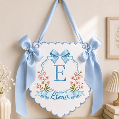 May include: A white scalloped wall hanging banner featuring a large blue embroidered letter E, pink floral accents, and the name Elena in blue script. The banner is decorated with two light blue satin bows and hangs from a matching blue ribbon.
