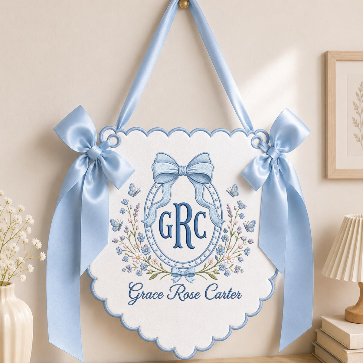 May include: A white scalloped wall banner featuring blue embroidery of the monogram GRC inside an oval frame, topped with a large blue bow. The design includes floral accents, butterflies, and the name Grace Rose Carter in blue script.
