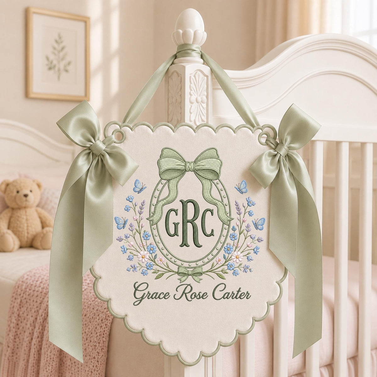 May include: A cream-colored scalloped wall hanging featuring embroidered sage green bows, blue butterflies, and floral accents. The center displays the monogram GRC inside an oval frame, with the name Grace Rose Carter embroidered below in dark green script.
