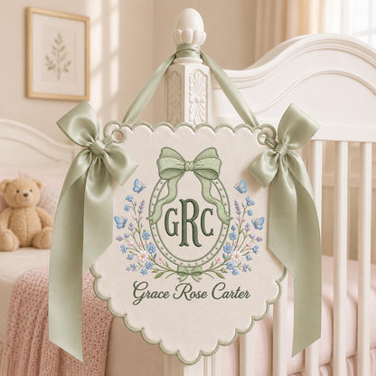 May include: A cream-colored scalloped wall hanging featuring embroidered sage green bows, blue butterflies, and floral accents. The center displays the monogram GRC inside an oval frame, with the name Grace Rose Carter embroidered below in dark green script.